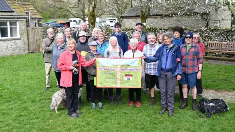 LDRS Nick Burton stands in a group of 19 people at the Pennine Way 2025 anniversary celebrations in Malham in the Yorkshire Dales. The group holds a green banner depicting Lancashire connections to the Pennine Way.