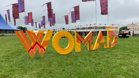 WOMAD sign at the festival grounds. The abbreviation is spelled out with large, orange and yellow printed letters on grass. The festival site can be seen behind in the distance - there are colourful flags, tents and stands. 