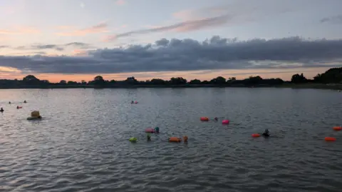 A dappled lake in the grey light of dawn. The heads of several swimmers in swimming caps can be seen, accompanied by pink and orange tow floats. The sky is a pinky blue.