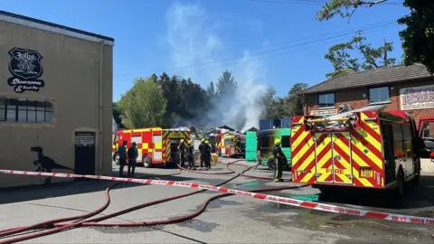 Red and white police tape stretching across a road. There are three fire engines and a green van which appears to be burned. There are firefighters around. There is light grey smoke is. the sky. The sky is blue.