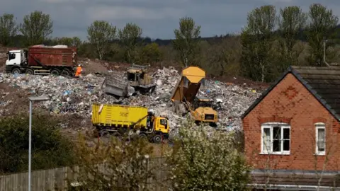 Reuters Three trucks emptying tonnes of rubbish at a landfill site on a hillside behind a house. There is a plough in the mound of rubbish alongside them.