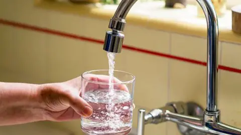 The image shows a close‑up of a person holding a clear glass under a kitchen faucet while fresh water flows into it. The hand is positioned securely around the glass, which is already partially filled. The faucet is metallic and curved, and the water stream appears clean and steady. In the background, the kitchen counter and tiled backsplash are slightly out of focus.