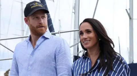 Reuters Harry is wearing a baby blue linen shirt and a black 'Invictus Australia' baseball cap. Meghan is wearing a darker blue and white striped shirt.