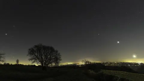 Josh Dury A wide-angle shot of the sky over the Mendip Hills in Somerset taken at night. The sky is dark and various stars are seen brightly shining with trees silhouetted in the foreground and a light haze over Bristol in the distance