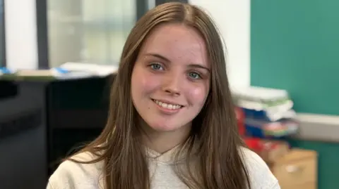 Ebony smiles at the camera. She has long brown hair and is wearing a white hoody. There is a piano behind her.