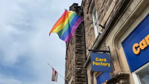 A Pride flag next to an St George's Cross flag above a Card Factory.
