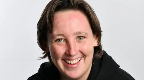 BBC Mhairi Black, a young woman with short loose brown hair, smiles into the camera against a grey backdrop. She is wearing a black hoody.