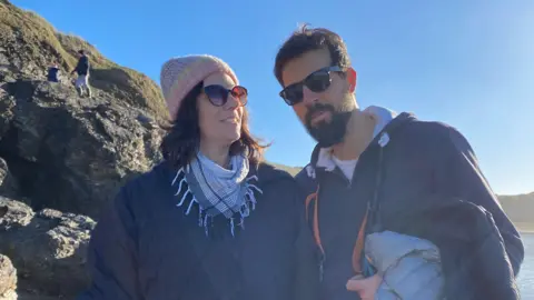 Karina Busquets Karina Busquets, left, is wearing a coat, scarf, hat and coat, and is stood on a beah with her husband. There are rocks to the left of her and see in the background