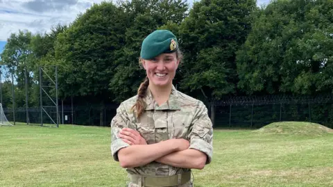 Lt Lily-Mae Fisher Lt Fisher stood in a field with her hands crossed in front of her chest. She is wearing a green beret. 