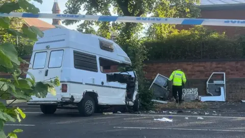 A crashed camper van in situ behind a police cordon with an officer in high vis nearby