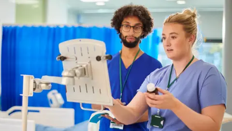 Getty Images Two hospital workers dressed in blue scrubs looking at what appears to be a clipboard. The female worker, who has pulled-backed blonde hair, is holding a bottle of medication. The male worker who has curly dark hair and a beard looks on.