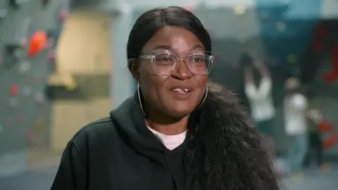 BBC A woman is smiling as she talks to the camera while being interviewed. She is wearing a black top. She has long black hair which is tied back and is wearing glasses. A blurred out climbing wall with people climbing on it can be seen in the background.