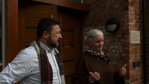 A man with a black hair and beard in a striped shirt and scarf is standing outside a wooden door to a brick building. Standing beside him is a woman with white hair. She is wearing a brown top, scarf and is holding her thumbs out.