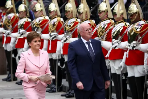 PA Media Nicola Sturgeon and Peter Murrell walk in front of a line of servicemen dressed in elaborate regalia. Sturgeon is wearing a pink suit. Murrell is in a navy blue suit and tie. 