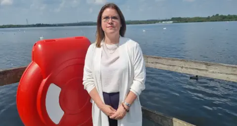 Staffordshire County Council A woman with dark hair past her shoulders stands in front of a body of blue water. A fence bar can be seen behind her, grey, with the rest plastic of a life ring to her right. She wears transparent-rimmed glasses and a white top over a white top. On the water in the distance, swans can be seen.