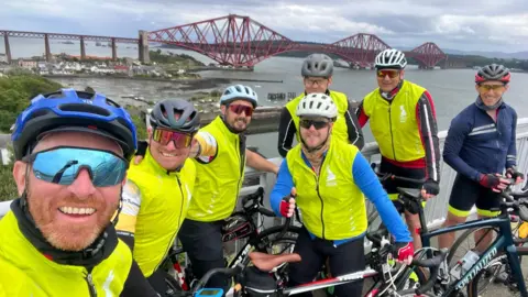 Jo Boswell A group of seven men wearing high-visibility jackets smiling at the camera with their bikes, with the Forth Bridge in the background.