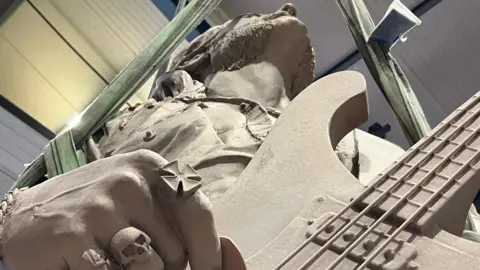A statue of a man holding a guitar, viewed from below and looking straight up to his chin. The fingers he is using to play the guitar have rings on them, including one of a skull.