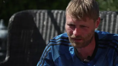 A man with blonde hair and a bear, wearing a blue football shirt, wears a serious expression as he sits on a wicker chair in a garden.