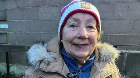 An older woman wearing a blue and red knitted hat and beige coat with a furry trim smiles while standing in front of a grey stone wall.