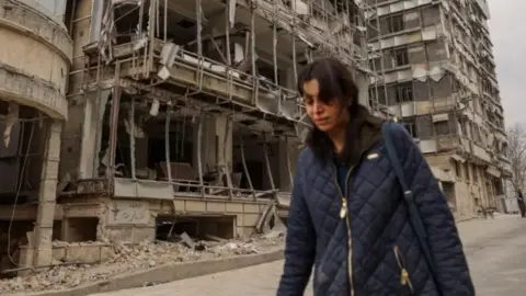 The image shows a heavily damaged multi‑storey building with its exterior walls torn open and debris scattered across the ground, with exposed concrete, broken windows, and hanging metal framework. In the foreground a woman wearing a quilted jacket and carrying a shoulder bag is walking past the ruined structure. 