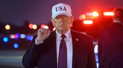 Close up of Donald Trump with his fist in the air after landing at Palm Beach international airport. It is night time and he is wearing a white cap with USA on it.
