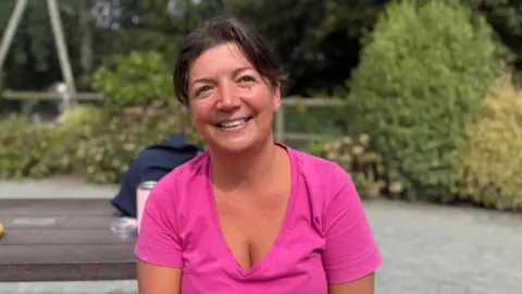 BBC A woman in her 30s wearing a pink top. She is sat on a bench and her hair is scraped back 