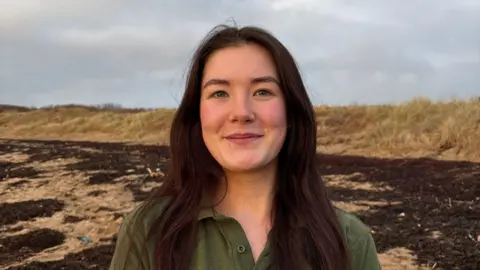 Caitlin Ganley with long brown hair stands on a beach smiling at the camera. She is wearing a green polo shirt.
