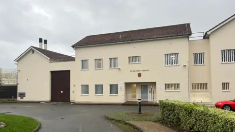 Les Nicolles Prison - A yellow building with the words Guernsey Prison on the walls. There are white bares on the windows and a grey door into the building. 