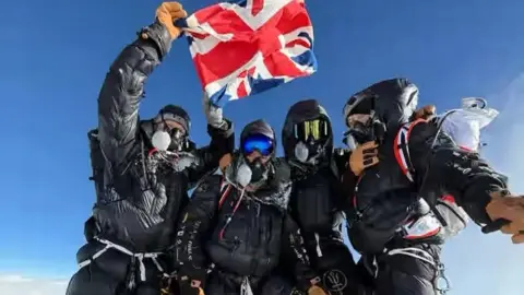 Four climbers standing together, dressed in climbing gear and masks, with one holding up a Union Jack flag 