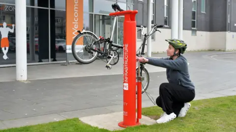 Hayley Crawford, customer service assistant at East Riding Leisure Bridlington, using the new cycle repair station. A large red metal post holds up a bicycle in position for repair. 