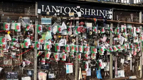A wall covered in pictures and a string of Welsh flags. At the top a board reads Ianto's Shrine in white writing. The pictures are attached to a rusty metal grid.