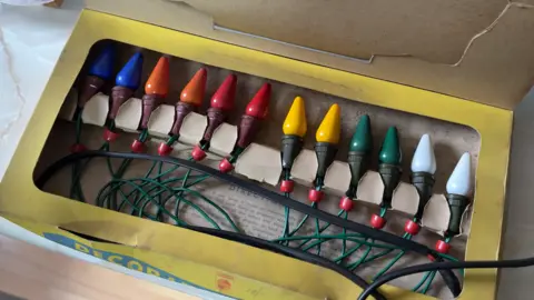 BBC / Grace McGrory The photo shows an old, yellow cardboard box which is open. It's filled with a multi-coloured string of lights - with each bulb painted either blue, orange, red, yellow, green or white. They are all connected to one another with a thin green wire. 