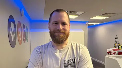 Paul Sewry smiling at the camera and stood inside the new centre. He has a large beard and is wearing a grey top. On the white wall to his left there are circle signs with different logos inside.
