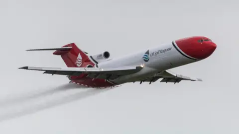 Julian P Guffogg / Geograph A Boeing 727 tri-jet flying on a grey day. It is painted with red and white livery. It is spraying a liquid from red and white booms attached to the back of the aircraft.