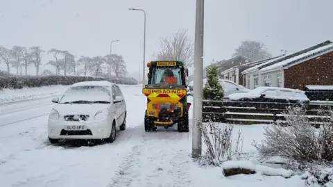 A silver car covered with snow is parked at the side of the road in Balerno. A grit spreading tractor is next to it on the pavement. It is yellow with a red and yellow banner and the word "spreading" in red text. A driver is visible in the cabin wearing an orange hi-vis vest. The ground, including the road, is almost completely covered in snow. The sky is grey and the trees in the background and foreground are bare.