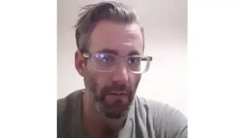 Avon and Somerset Police Image of Michael Wheeler wearing glasses. He is wearing a grey t-shirt and has a beard.