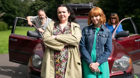 Two Brothers Productions Ashley in a scene from Dinosaur. She is standing wearing a flowery dress and beige coat, next to the actress Kat Ronney, who is wearing a denim jacket and turquoise dress. They are standing in front of a maroon car, with the characters of Nina's parents standing either side of it and looking at them.