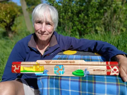 Lesley Sleight Lesley Sleight, wearing a blue jumper, leans on a table where the baton rests. It is a three-sided wooden baton, featuring coloured paintings of Manx heritage sites, and cultural events. 