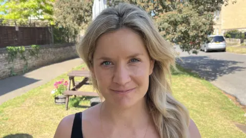 Francesca Martin is standing in a garden which is next to a road and a pathway. She has long blonde hair and is wearing a black short sleeved T-shirt. There is a bench behind her which has a parasol. 
