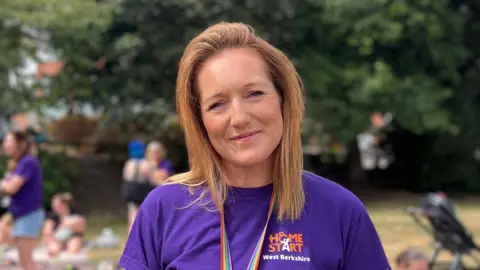 Grace is wearing a purple t-shirt with home start west berkshire logo. She is behind people sitting on picnic blankets with their children. The background is blurred. 