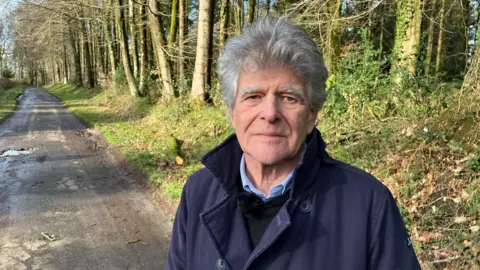 A man with bushy grey hair, a blue coat and a blue jumper is standing on a country road lined by trees with grass verges either side and potholes visible in the road surface behind him