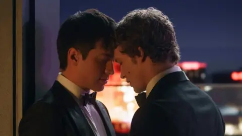 PA Media Two men wearing tuxedos with their heads touching on a balcony with the night sky and lit buildings behind them.
