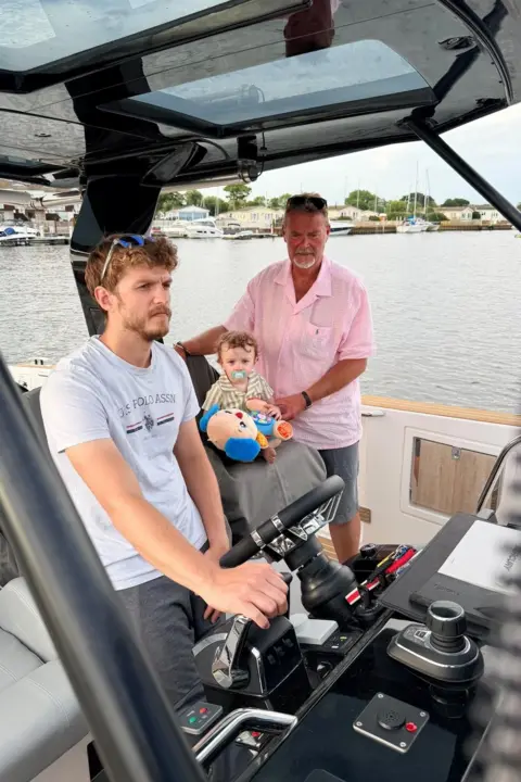 Andy Golding Max Golding stands at the helm of a motor boat in Christchurch Harbour while Andrew Golding and Max's son who is a young child, sit beside the controls. Boats and waterside properties can be seen in the background.