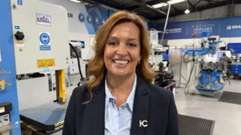 A woman with long curly brown hair stands in the same factory environment wearing a navy blue blazer and light blue unbuttoned shirt.