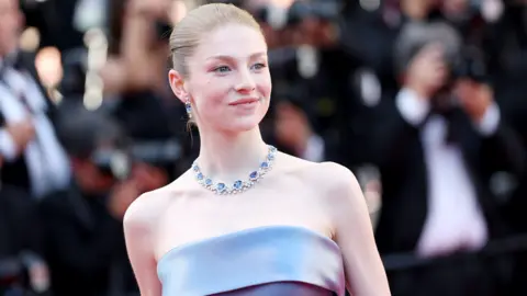 Hunter Schafer at a red carpet event wearing a blue dress and matching necklace and earrings.