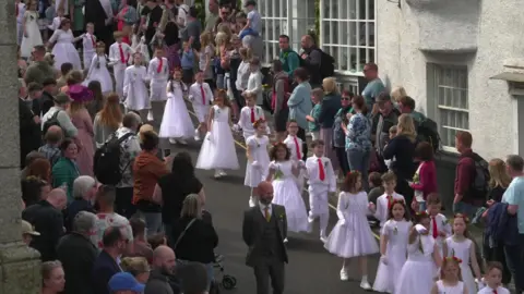 A procession of children dressed in white dance through the streets of Helston, watched by a crowd of happy people 