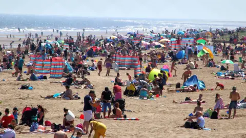 Keith Mayhew/SOPA/Getty A crowded beach can be seen filled with people lying on a golden, sandy beach, playing in the sand and enjoying the sun. Blue, red and white wind breakers dot the scene. The sea is blue.