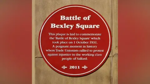 A red plaque on a brown stone background, reads in white writing: Battle of Bexley Square. This plaque is laid to commemorate the ' Battle of Bexley Square' which took place on 1 October 1931. 