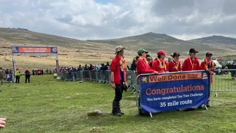 BBC The team stood in front of a banner with the words "Well Done" and "Congratulations" below. The sign is orange and blue. The team is stood behind the barrier and looking to the right. There's crowds of people behind them stood at the finish line. 