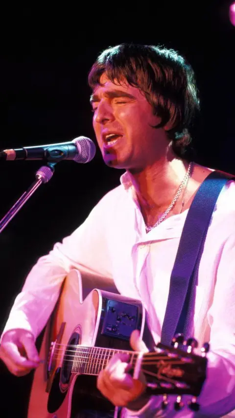 Getty Images Noel Gallagher playing a guitar and signing on stage next to a microphone in a stand. He is wearing a white long-sleeved top.